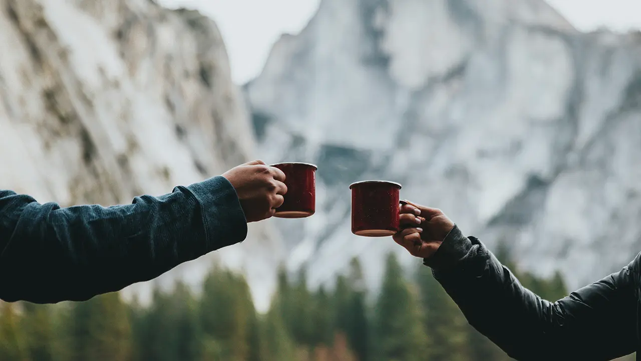 Vor dem Hintergrund einer Bergkulisse stoßen zwei Menschen miit Kaffeebechern an.