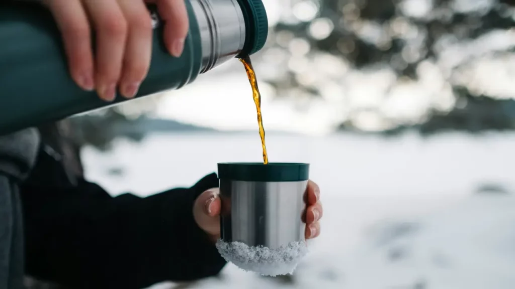 Kaffee wird aus einer Thermoskanne in einen Becher gegossen, Ein winterliche Landschaft im Hintergrund.