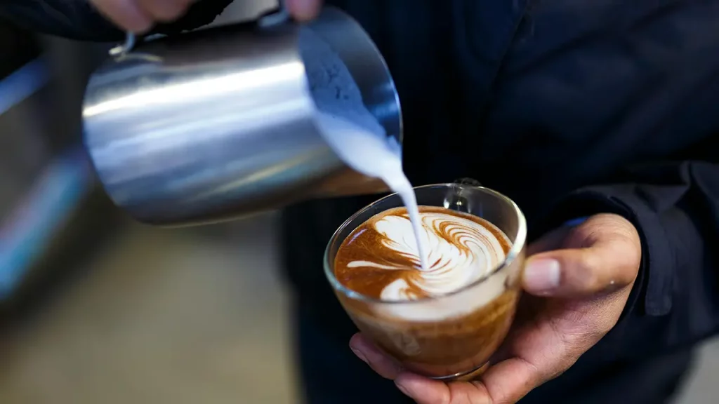 Nahaufnahme von Händen, die Latte Art zubereiten: Ein Barista gießt mit einem Edelstahlkännchen geschäumte Milch in eine Glas-Tasse Cappuccino und formt ein Herzmuster.